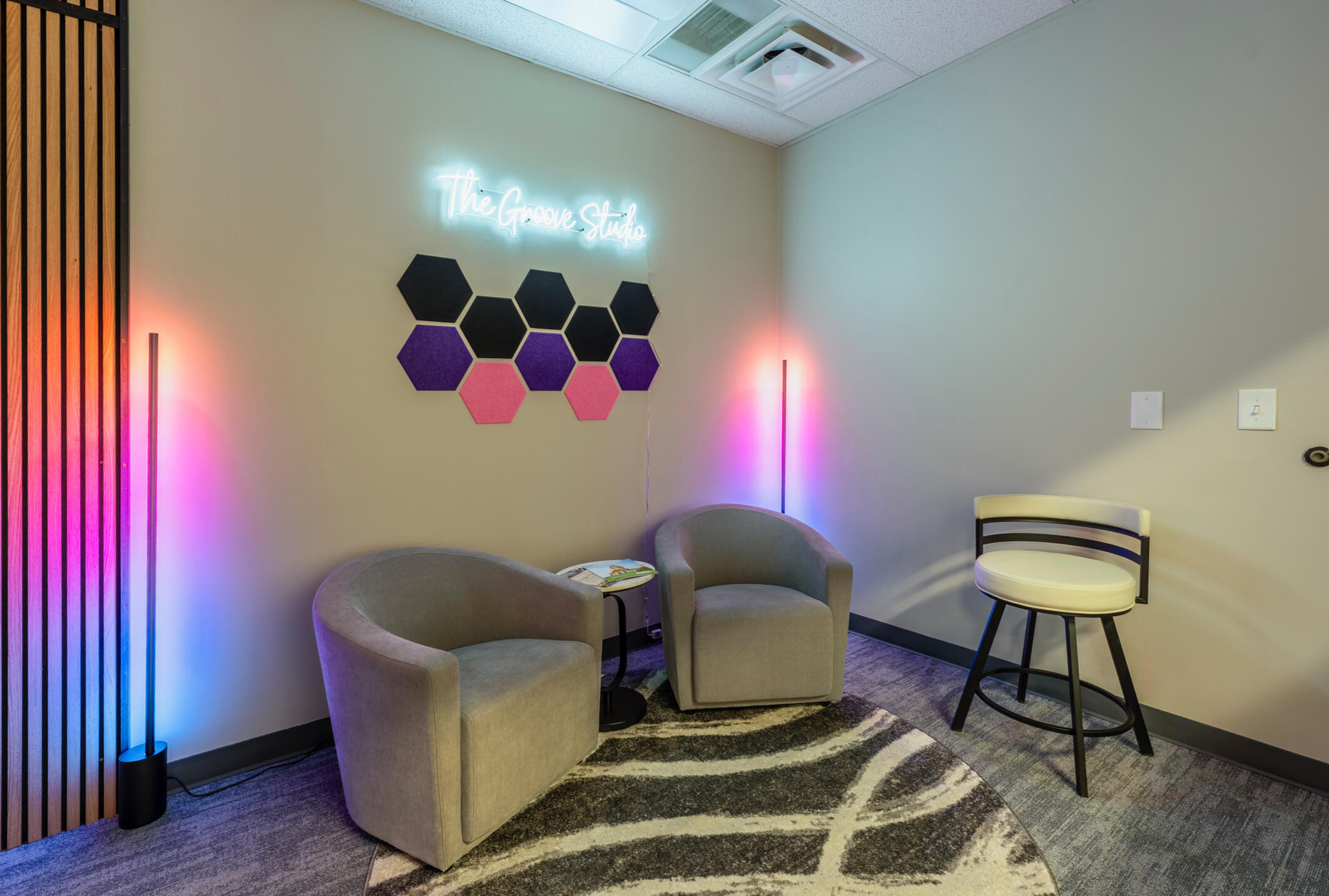 Interior view of a modern lounge area featuring white neon sign reading 'The Groove Studio' mounted on wall above hexagonal acoustic panels in black, purple, and pink colors, two beige upholstered armchairs, side table with magazines, bar stool, and pink-p
