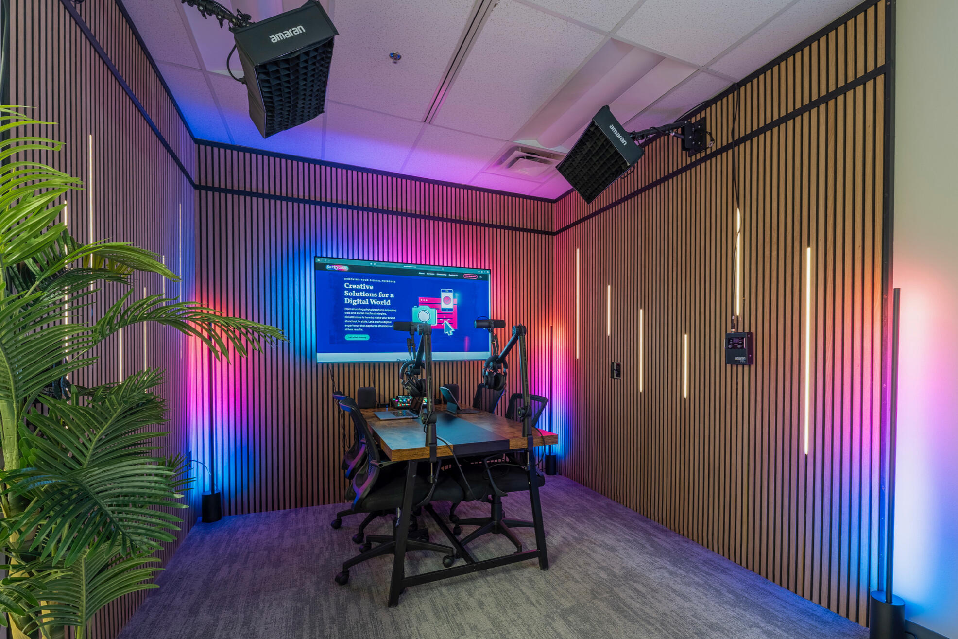 Wide-angle view of a professional podcast recording studio with vertical wood acoustic panels covering walls, gradient LED lighting transitioning from blue to pink to orange, large wall-mounted display screen, wooden recording table with two professional m